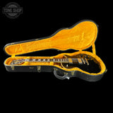 A black electric guitar shown inside an open hard case with bright yellow-orange plush lining, photographed against a solid black background. A Tone Shop watermark appears in the upper left corner.