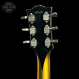 Close-up of the headstock of a vintage electric guitar, showing six silver tuning pegs on a black backplate with a gold body visible below, set against a solid black background.