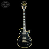 A black Gibson Les Paul Custom electric guitar with gold hardware and white block inlays, shown against a solid black background with a Tone Shop watermark in the top-left corner.