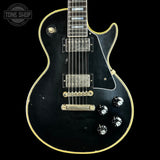 A black electric guitar with gold trim and hardware, photographed against a solid black background. A circular 'TONE SHOP' watermark is visible in the top-left corner.