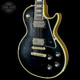 A black electric guitar with gold trim and gold hardware, photographed against a solid black background. A circular 'TONE SHOP' watermark is visible in the top-left corner.