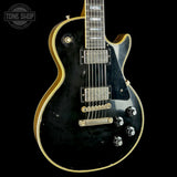 A black electric guitar with gold trim, featuring a glossy finish, dual humbucker pickups, and chrome hardware, shown against a solid black background with a Tone Shop watermark in the top left corner.