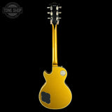 A gold-finish electric guitar shown from the back, featuring a black tailpiece, silver tuning pegs, and a circular gold badge on the body, set against a solid black background.