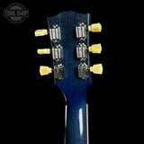 Close-up of the headstock of a blue electric guitar against a black background, showing six gold tuning pegs and a Tone Shop watermark in the upper left corner.