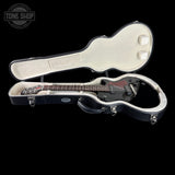 A black electric guitar displayed inside an open hard case with a cream-colored interior, set against a solid black background. A Tone Shop watermark is visible in the top-left corner.