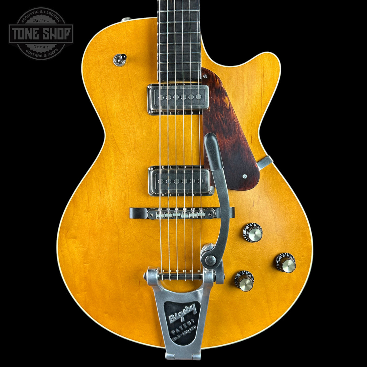 A close-up of a yellow-orange electric guitar body with a dark brown pickguard, chrome hardware, and a silver Leslie speaker mount against a black background. A Tone Shop watermark is visible in the top left corner.