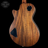 The back of a double-cutaway electric guitar with a rich brown wood grain finish, featuring black hardware and a circular control hole, shown against a solid black background with a Tone Shop watermark in the top left corner.