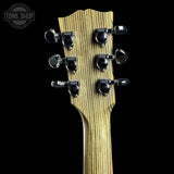 Close-up of the headstock of a guitar with a natural wood grain finish and six black tuning pegs, shown against a solid black background. A Tone Shop watermark is visible in the top left corner.