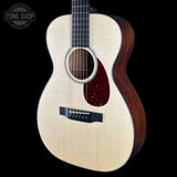 An acoustic guitar with a natural wood top, dark reddish-brown sides, and a black pickguard, shown against a solid black background with a Tone Shop logo in the top-left corner.
