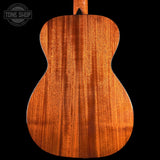 Close-up of the back of a wooden acoustic guitar showing the rich grain of the mahogany wood, set against a solid black background with a Tone Shop watermark in the top left corner.