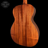 Close-up of the back of an acoustic guitar showing a rich, warm reddish-brown wood grain finish, set against a solid black background with a Tone Shop watermark in the top left corner.