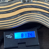 Close-up of a guitar body with wavy wood grain stripes above a digital smart weigh scale displaying 8.134 oz on a dark wooden surface.