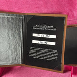 An open Gibson Custom Certificate of Authenticity booklet showing a black card with handwritten serial numbers on a pink textured fabric background.