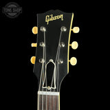 Close-up of a Gibson acoustic guitar headstock in black with gold lettering and gold-toned tuning pegs and truss rod covers, shown against a black background with a Tone Shop watermark in the upper left.