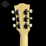 Close-up of the headstock of a guitar with a cream-colored backplate and six black tuning pegs with white tuning keys, shown against a solid black background.