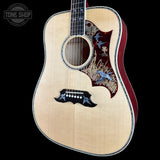 A close-up of an acoustic guitar body against a black background, featuring a natural wood top, a dark red double-cutaway pickguard with blue bird and grass inlay, and a black bridge.