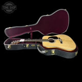 An acoustic guitar with a natural wood body and dark neck, displayed inside an open black hard case with a burgundy interior. The Tone Shop logo is visible in the top-left corner.