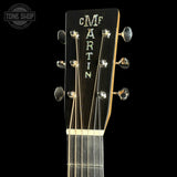 Close-up of the headstock of a Martin acoustic guitar against a black background, showing the brand name 'CMF MARTIN' in gold lettering on a dark wood headstock with silver tuning pegs.