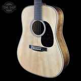 A close-up studio shot of an acoustic guitar body against a black background, showing a natural wood top, dark pickguard, and part of the neck. A Tone Shop watermark is visible in the upper left corner.