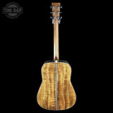The back of an acoustic guitar with a highly figured wood top, showing a prominent grain pattern in warm golden-brown and tan tones, photographed against a solid black background.