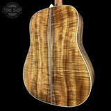 The back of an acoustic guitar featuring a rich, figured wood grain top in warm golden-brown tones, with white binding, shown against a solid black background. A Tone Shop watermark is visible in the top-left corner.