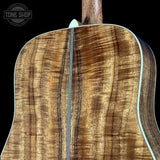 Close-up of the back of an acoustic guitar showing a rich, figured wood grain finish with a black background and a Tune Shop watermark in the top left corner.