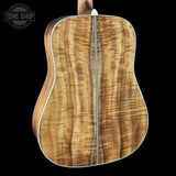 The back of a wooden acoustic guitar with a prominent figured holly pattern in warm brown and golden tones, set against a solid black background with a Tone Shop watermark in the top-left corner.