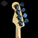 Close-up of the headstock of a bass guitar with a natural wood finish and four black tuning machines, shown against a solid black background. A Tone Shop watermark is visible in the top left corner.