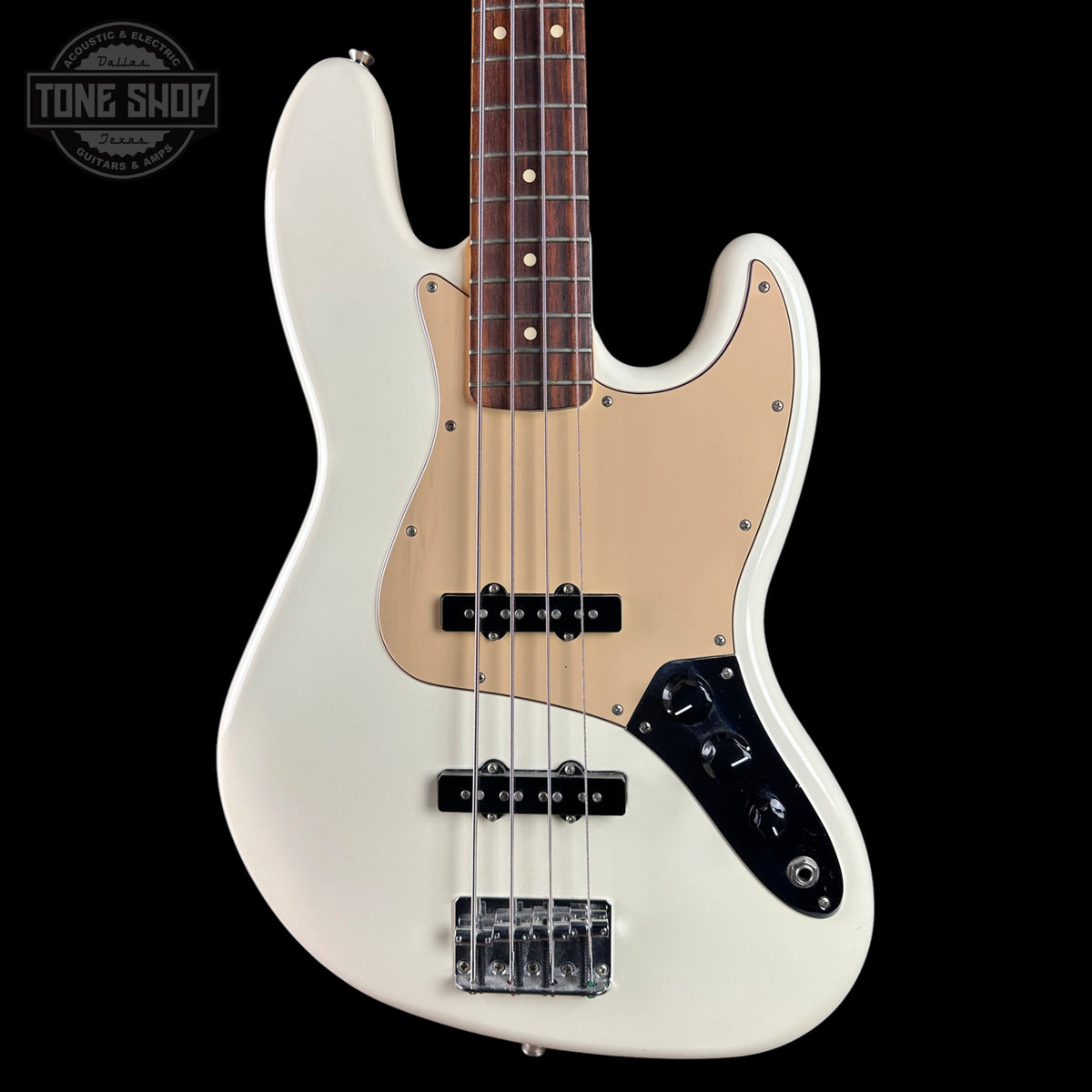 A white electric bass guitar with a beige pickguard and dark brown neck, shown against a solid black background. A Tone Shop logo is visible in the top-left corner.
