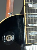 Close-up of a black electric guitar body showing the tailpiece area, fretboard, strings, and a round black control labeled RHYTHM TREBLE.