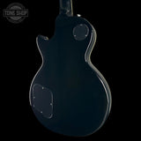 The back view of a glossy black electric guitar against a solid black background, featuring a circular Tone Shop watermark in the upper left corner.