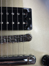 Close-up of a guitar's bridge and pickups, showing a black humbucker pickup, a chrome tremolo system, and part of a metallic gold or champagne-colored guitar body.