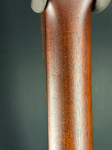Close-up of a polished brown wooden rifle stock with visible wood grain and a small portion of a light-colored grip at the left edge, set against a dark gray background.