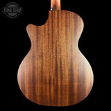 Close-up of the back of an acoustic guitar showing a rich, warm brown wood grain finish on a solid black background, with a Tone Shop watermark in the top left corner.