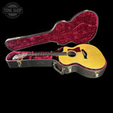 An acoustic guitar with a natural wood top and dark pickguard displayed inside an open hard case with a deep burgundy velvet lining, set against a solid black background.
