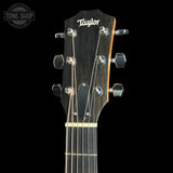 Close-up of the headstock of a Taylor acoustic guitar against a black background, showing the brand name 'Taylor' in white script on a dark wood headstock with six tuning pegs.