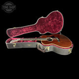 An acoustic guitar with a brown wooden body and black headstock, displayed inside an open gray hard case with a deep red velvet interior, set against a solid black background.