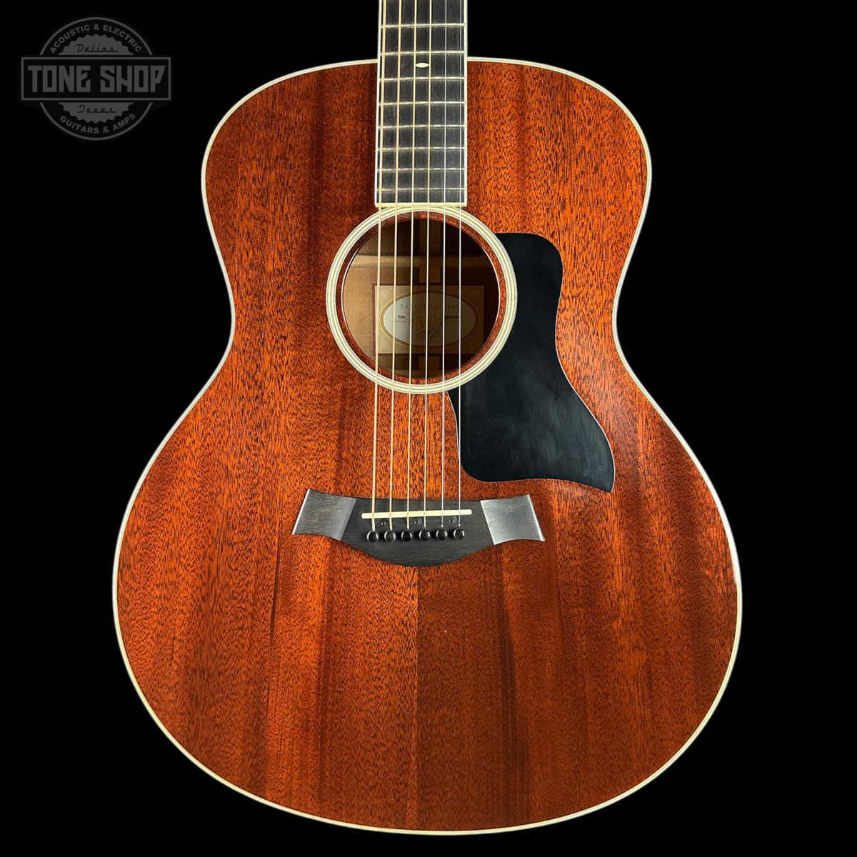 A close-up studio shot of an acoustic guitar body with a warm reddish-brown wood finish, black pickguard, and white binding, shown against a black background. A Tone Shop watermark is visible in the upper left corner.