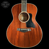 A close-up studio shot of an acoustic guitar body with a warm reddish-brown wood finish, black pickguard, and white binding, shown against a black background. A Tone Shop watermark is visible in the upper left corner.