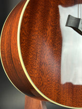 Close-up detail of an acoustic guitar's richly carved wood backplate, showcasing a deep reddish-brown grain pattern and a gold-toned binding edge.