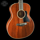 A close-up of a Taylor acoustic guitar body with a rich reddish-brown wood finish, featuring a black pickguard and white binding, set against a solid black background with a Tone Shop watermark in the top left corner.