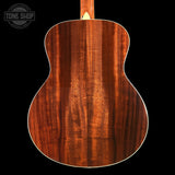 Close-up of the back of an acoustic guitar showing a rich, warm brown wood grain finish, set against a solid black background with a Tone Shop watermark in the top left corner.