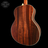 The back of an acoustic guitar with a rich, warm brown wood grain finish and a white binding edge, shown against a solid black background with a Tone Shop watermark in the top left.
