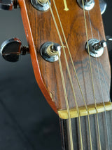 Close-up of an acoustic guitar headstock showing six tuning pegs, gold strings, and a warm brown wood finish against a dark background.