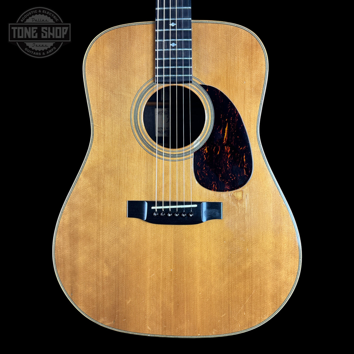 A classic acoustic guitar with a natural wood finish and a dark tortoiseshell pickguard, shown against a solid black background with a Tone Shop watermark in the top left corner.