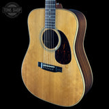 An acoustic guitar with a natural wood body and dark brown sides, shown against a solid black background. A circular 'TONE SHOP' watermark is visible in the top-left corner.