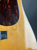 Close-up detail of an acoustic guitar body showing the natural wood top, a dark tortoiseshell pickguard, and part of the sound hole cover.