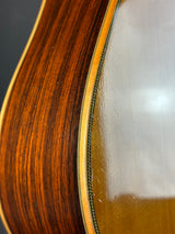 Close-up detail of an acoustic guitar body showing the rich brown wood grain on the back, the glossy varnished front, and a decorative green-and-white braid along the edge.