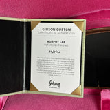 A Gibson Custom Certificate of Authenticity insert for a Murphy Lab Ultra Light Aging instrument, displayed inside an open gold-lined presentation case on a pink velvet background.