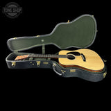 An acoustic guitar with a natural wood body and dark neck displayed inside an open black hard case with a green felt lining, set against a solid black background. A 'TONE SHOP' logo watermark is visible in the top-left corner.